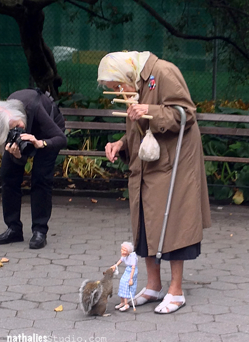 Older woman in a park with a marionette of herself that feeds a squirrel. A women next to her taking a photo of the scene